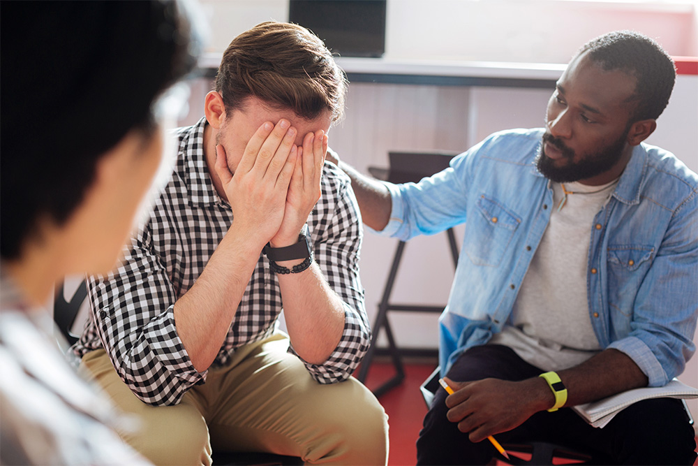 A distressed man sits with his face in his hands, while another person offers support by gently placing a hand on his shoulder. This image represents the compassionate crisis care provided by Agency for Community Treatment Services, Inc. (ACTS), where individuals facing emotional or mental health challenges receive immediate support and understanding in a safe environment.