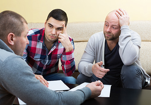 Three men sit together in serious conversation, with two of them appearing distressed and concerned. This image represents the crisis intervention services provided by Agency for Community Treatment Services, Inc. (ACTS) for adults experiencing crises due to intoxication or drug impairment, offering immediate support and guidance during critical moments.