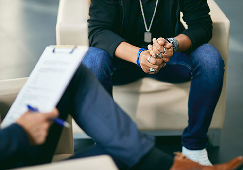 A person sits with hands clasped during a counseling session, while a therapist takes notes on a clipboard. This image represents the outpatient treatment services provided by Agency for Community Treatment Services, Inc. (ACTS), offering individuals professional support and guidance on their recovery journey in a non-residential setting.