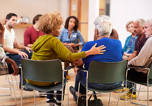 A group of people sit in a circle during a support meeting, with one woman gently comforting another by placing a hand on her back. This image represents the substance abuse support services offered by Agency for Community Treatment Services, Inc. (ACTS), providing compassionate care and group therapy for individuals on their recovery journey.