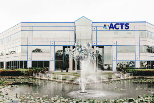 A large modern office building with an ACTS sign displayed prominently on the facade. The building has a symmetrical design with large glass windows and a central glass atrium. In front of the building, there is a pond with a fountain spraying water into the air, surrounded by greenery and water lilies.