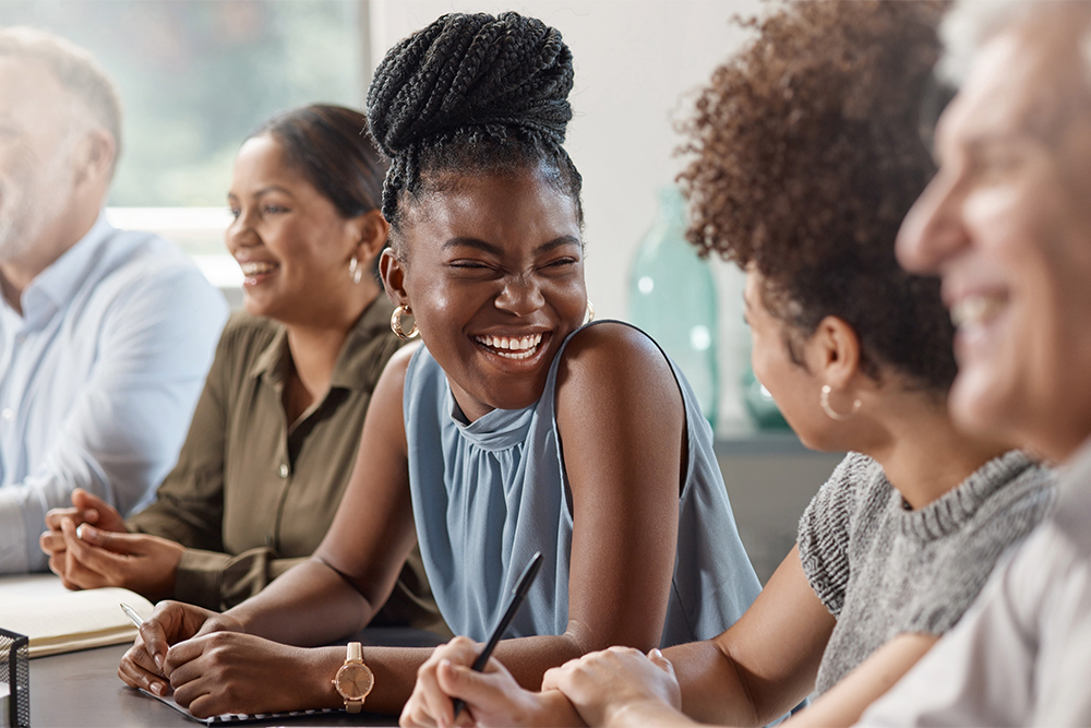 A group of people sitting at a table, laughing and smiling during a conversation. The focus is on a young woman with braided hair in a light blue sleeveless top, who is laughing joyfully while holding a pen. Around her, other individuals, including a woman with curly hair and people in the background, are also smiling and engaged.