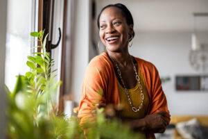 A smiling woman with dark hair tied back is standing near a window, looking outside. She is wearing a mustard yellow top, an orange cardigan, and a long necklace, with large hoop earrings. Green plants are in the foreground, and the background shows a bright, modern interior space with natural light.