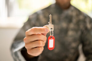 A person in a camouflage military uniform holding a silver key with a red key tag close to the camera. The person's hand is in focus, while their face and uniform are blurred in the background.