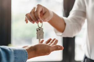 A close-up of two hands exchanging a house-shaped keychain with a key. One person, wearing a white shirt, is handing over the key to another person, whose open hand is reaching to receive it. The background is blurred, showing a bright window with natural light.