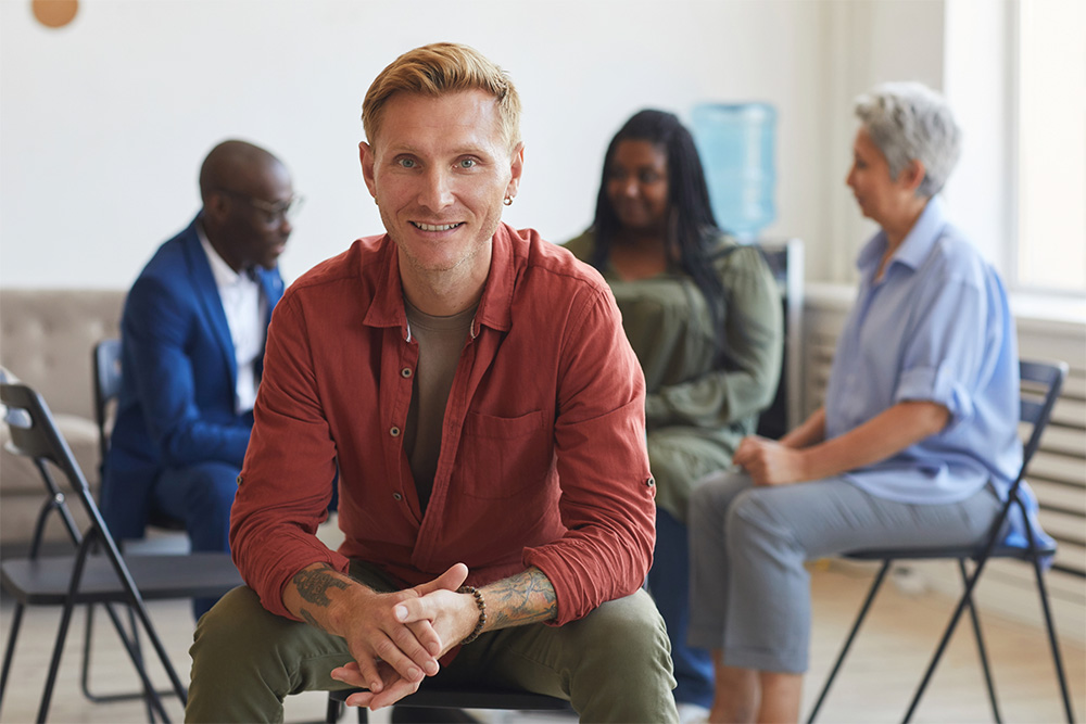 A man with short blond hair, tattoos on his arm, and wearing a red shirt is sitting and smiling at the camera in a bright room. Behind him, three people—a man in a suit, a woman in green, and another woman with gray hair in blue—are sitting on chairs, engaged in conversation.